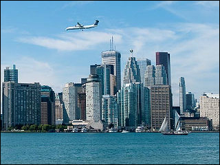 Photograph of the Toronto skyline seen from over the water, with a commercial jet in flight over the skyline and boats visible near the harbor.