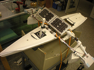A long slim white hull sits in a cradle, with PVC/Styrofoam pontoons extending to the sides, and a solar panel and periscope assembly sitting on top.