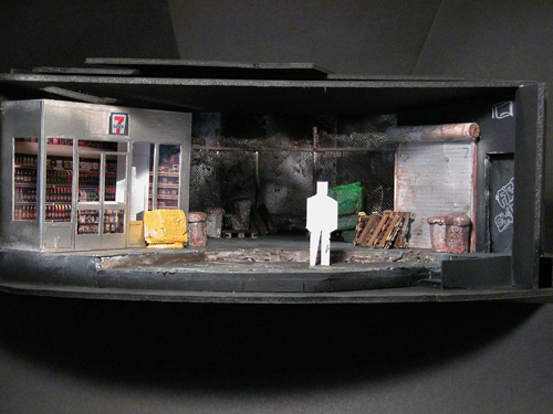 A diorama featuring a 7-11 on one side, some pallets and a sliding metal door on the other, chainlink fence along the back, and a paper stick figure center stage for scale.