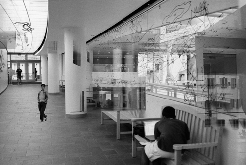 Photograph of hallway with art on ceiling and a glass wall; also people walking, sitting on a bench.
