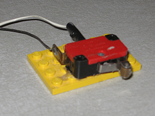 A red plastic box has a flexible metal arm extending from one side, with a plastic tab protruding from a slot in the box near the end of the arm.