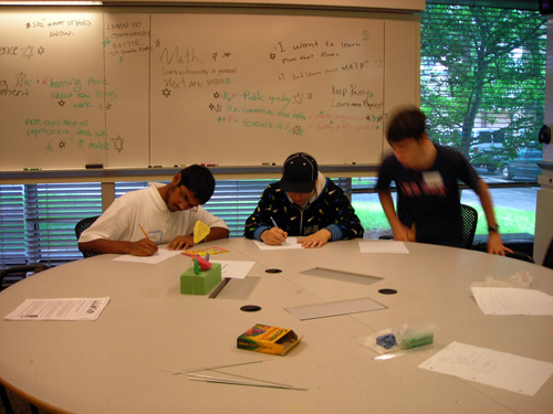 Three students sit at a table working on a project. A large green brick of modeling clay and crayons sit on the table.
