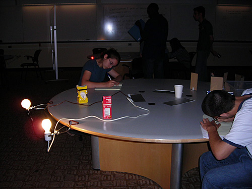 Two students sit at a table making observations about a pair of lit lightbulbs.