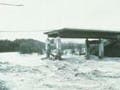 A photograph of a bridge failure in progress.  Half of the bridge has been washed away and the remaning structure appears to be crumbling.