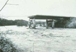 A photograph of a bridge failure in progress.  Half of the bridge has been washed away and the remaning structure appears to be crumbling.
