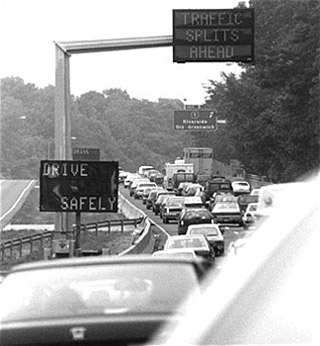 A black and white photograph of traffic congestion.  A seemingly endless line of cars sit on a highway underneath signs that read 'Drive Safely' and 'Traffic Splits Ahead'.