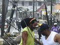 A photo of two men standing in debris after Hurricane Maria hit Puerto Rico. They are surrounded by damaged houses and fallen telephone poles and wires. 