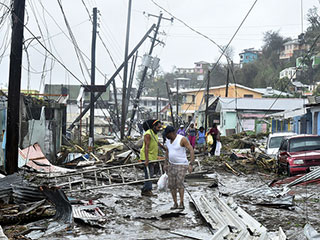 A photo of two men standing in debris after Hurricane Maria hit Puerto Rico. They are surrounded by damaged houses and fallen telephone poles and wires. 