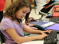A young girl in a lilac shirt sits at a computer working on homework.