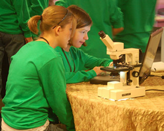 Two girls in green shirts use a laptop computer and microscope.