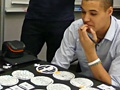 A student is seated at a table, looking down at a homemade board game. The game consists of a series of round spaces that the player must move through.