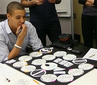 A student is seated at a table, looking down at a homemade board game. The game consists of a series of round spaces that the player must move through.