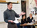 A teacher stands in a classroom with students seated and taking notes. The governor of Massachusetts sits with the students.