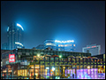 A photograph of a city at night time. There is a plaza in the foreground, a large glass building in the middle, and a city skyline in the background. Many of the buildings have neon lights of all colors. The lights are reflected off the glass building.