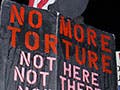 A protester stands with a black hood over his face while holding a sign that reads “No more torture, not here, not there, nowhere.”