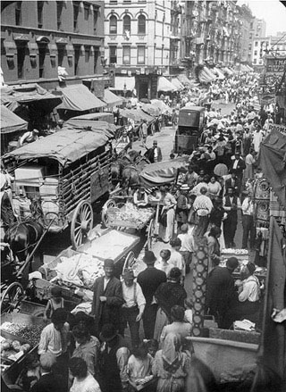 A black and white photo of an urban street crowded with people, wagons, carts, and horses.