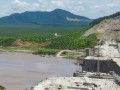A large, partially-completed dam in a broad river valley, with hills in the distance.