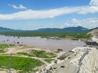 A large, partially-completed dam in a broad river valley, with hills in the distance.