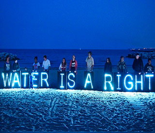 Protesters stand on a beach at nightfall with a neon blue sign that reads “Water is a right”.
