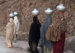 Pakistani women clad in traditional clothing carrying water in metalic vessels on their heads .