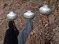 Pakistani women clad in traditional clothing carrying water in metalic vessels on their heads .