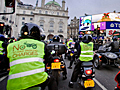 Photo of a large group of motorcyclists riding down a city street, some wearing vests with protest message 'No to bike parking charges!'