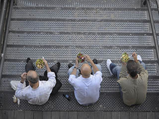 A photograph of three men sitting together on a staircase, eating salads.