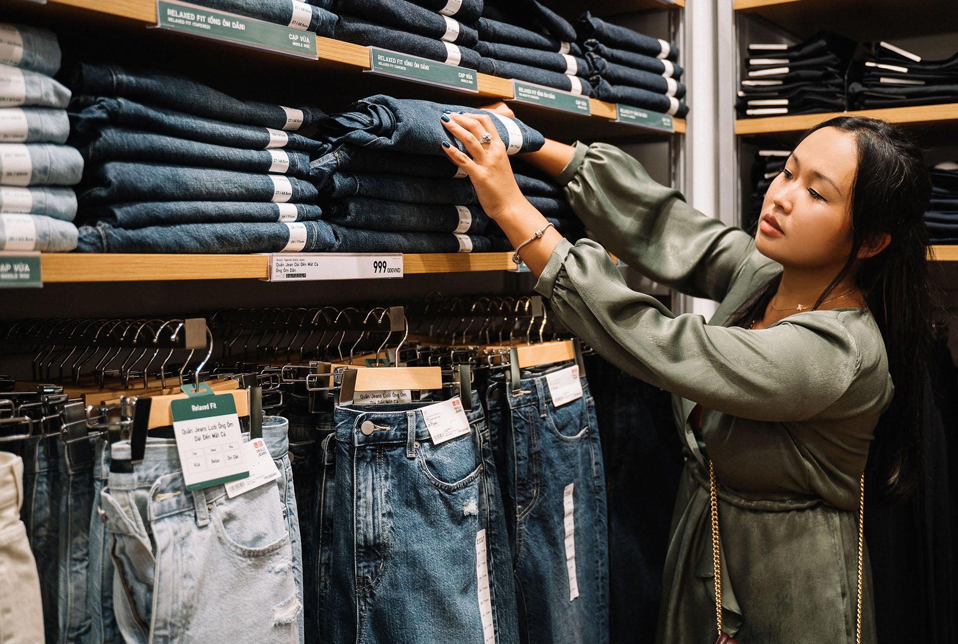 A woman in an olive green top is browsing a stack of dark wash denim jeans on a wooden shelf. 