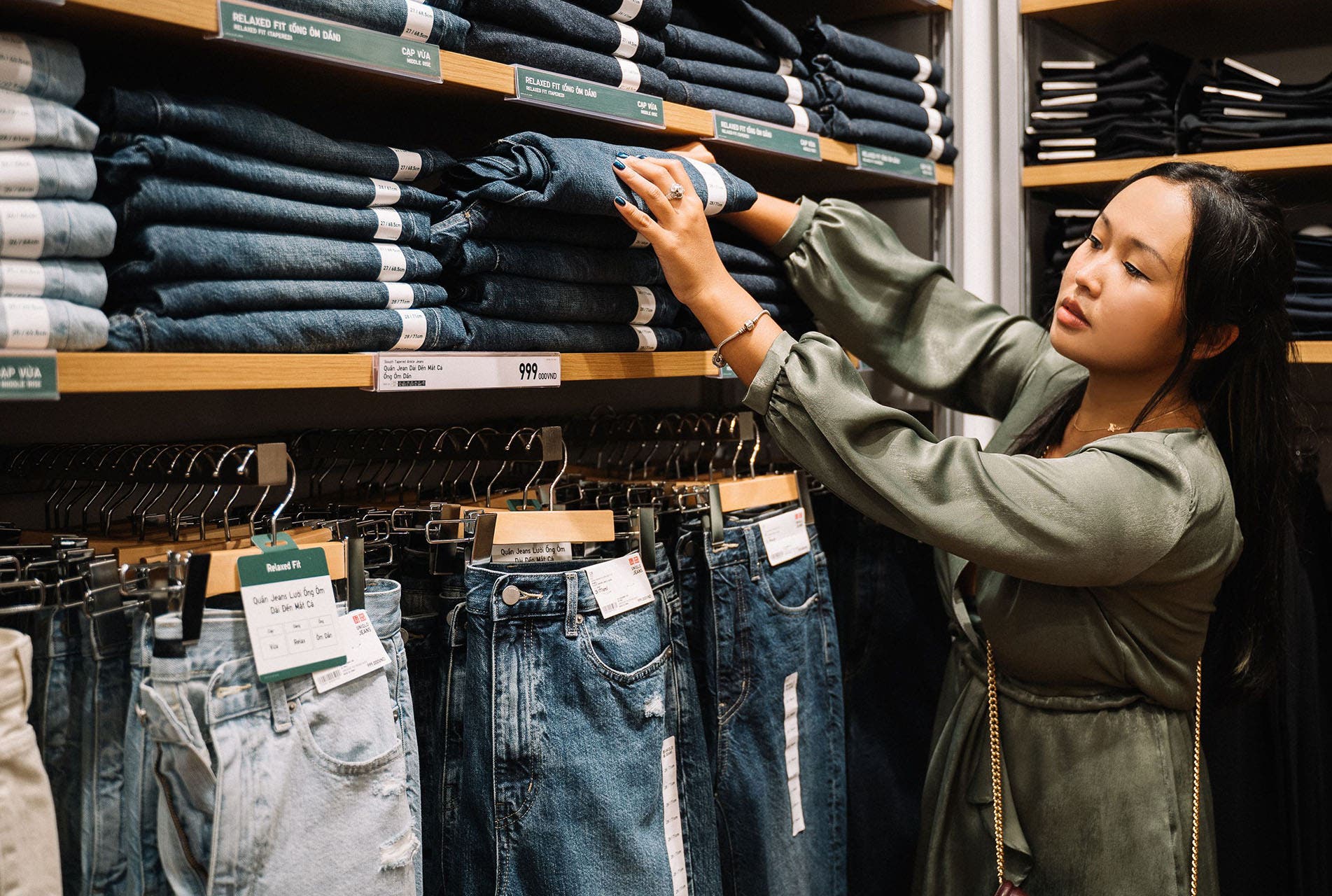 A woman in an olive green top is browsing a stack of dark wash denim jeans on a wooden shelf. 