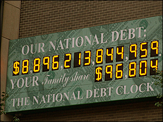 A photograph of a billboard sized electronic clock on a brick wall that displays the national debt of the United States.