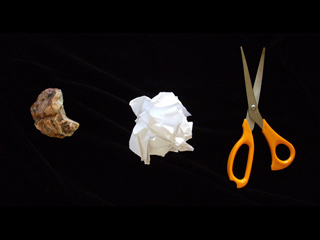 A rock, a crumpled piece of paper, and a pair of orange-handled scissors are set side-by-side on a black background.