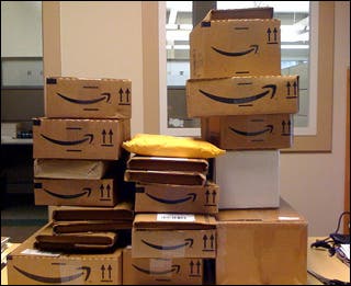 A dozen boxes from Amazon.com are stacked up on top of each other on a desk.