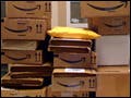 A dozen boxes from Amazon.com are stacked up on top of each other on a desk.