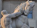 A photograph of a stone statue depicting a muscular and shirtless man pulling on the reins of an equally muscular horse.
