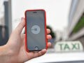 Someone holds a smartphone with an Uber app open on it. A taxi is seen in the background. 