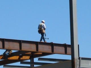 A construction worker, wearing a hard hat, walks along a steel I-beam on the upper part of the frame of an unfinished building.