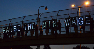 People stand on a highway overpass at night holding lighted signs that spell out "Raise the Min Wage."