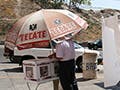 A man stands under a large umbrella, and places his votes into white boxes. A sign hangs nearby that says "el voto es libre y secreto."  