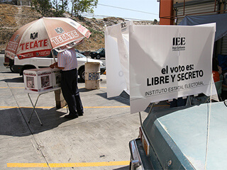 A man stands under a large umbrella, and places his votes into white boxes. A sign hangs nearby that says "el voto es libre y secreto."  