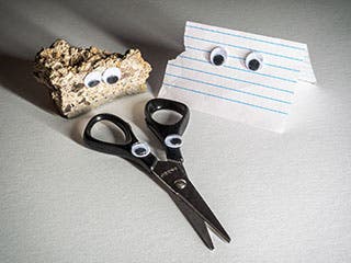 Photo of a beige-colored, bumpy rock, a piece of lined paper, and a pair of scissors, all with googly eyes attached to them.