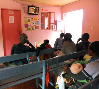 Rows of benches in a waiting room of a community clinic, women and children sit and watch television.