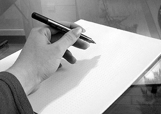 Black and white photo of a hand holding a pen, prepared to write on a sheet of paper.