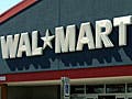 A small blue car is parked in front of a Wal-Mart entrance.