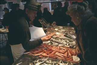 A fishmonger and a customer interacting across a counter covered with bins of fish in Venice's Rialto Markets.