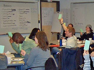 Students sitting in class raise their hands while holding green notecards.