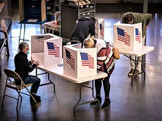 In an open room, people positioned behind dividers emblazoned with the U.S. flag fill out ballots.