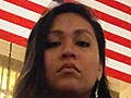 A solemn-faced brown-skinned woman, seated beneath the American flag, holds a sign with the text: Black Lives Matter.