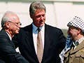 Two gray-haired gentleman, one wearing a suit, the other wearing a brown jacket and a long scarf, shake hands while a younger man stands between them.