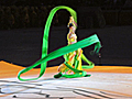 Dozens of people dressed in bright green uniforms carry poles that support a movable stage. A dancer twirling a green ribbon, stands on the stage.
