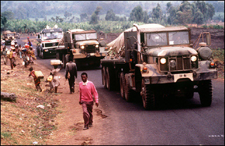 Three military vehicles pass alongside walking villagers on a narrow road.
